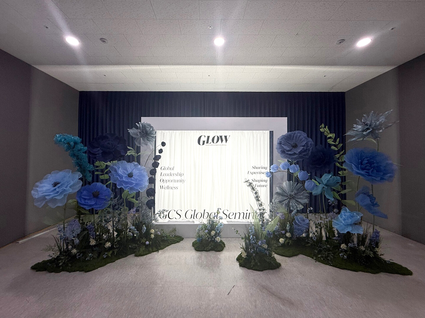 성남 센터엠 GLOW 세미나 행사장 무대 전경, 네이비 블루 방염 암막커튼 배경에 흰 현수막과 양옆 블루 플라워, 바닥의 그린 장식