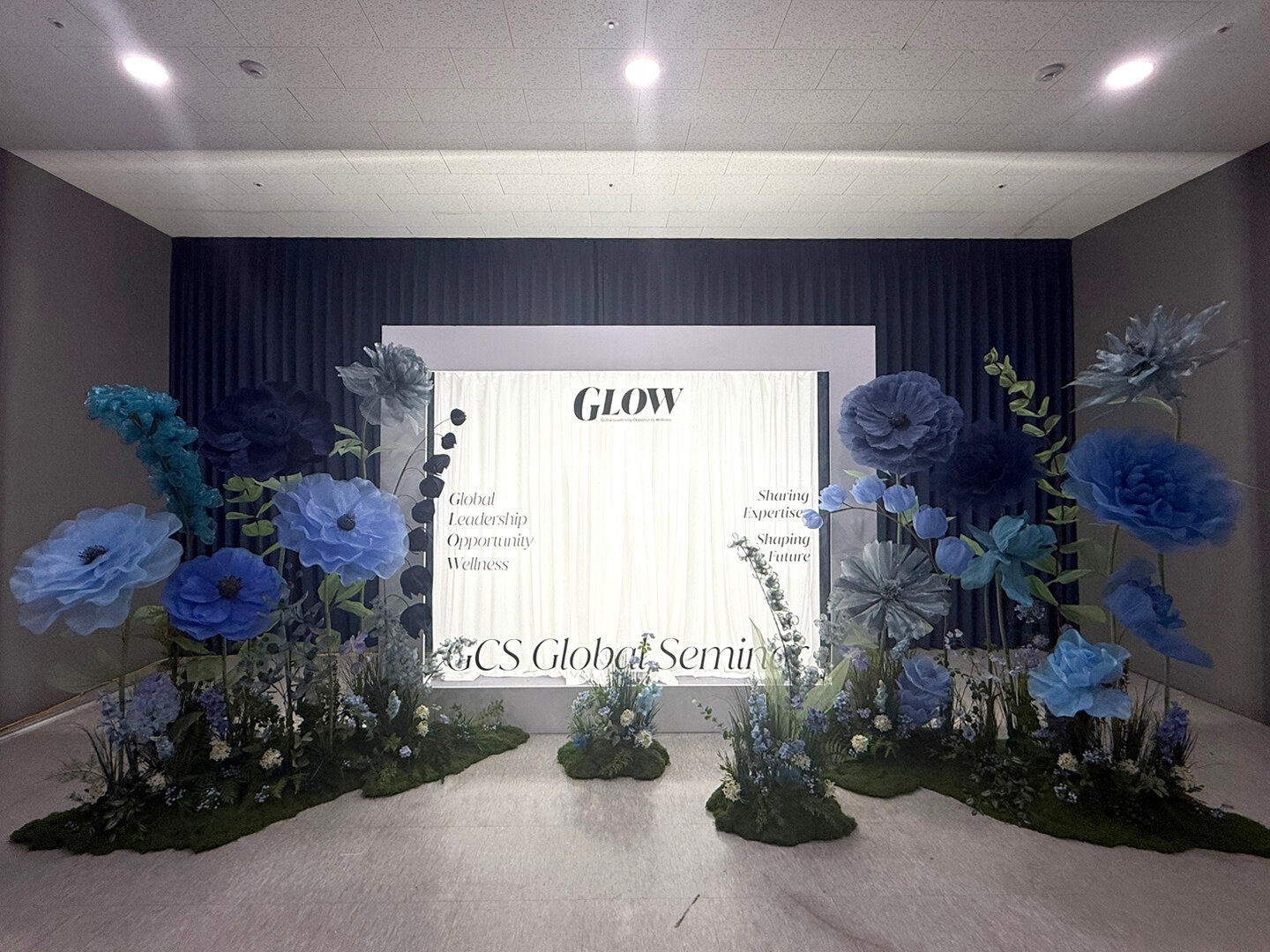 성남 센터엠 지식산업센터 GLOW 세미나 행사장, 벽 한 면 전체에 네이비 블루 방염 암막커튼을 바닥까지 치고 그 앞에 흰 세미나 현수막과 대형 블루 플라워 장식이 놓인 무대 배경